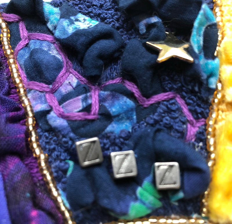 a close up of dark purple blue fabric and embroidery. running along the left side is a row of gold beads, upper right side a gold star. In the middle is an embroidered purple melatonin molecule shap and under that 3 little squares and each has a Z in it.